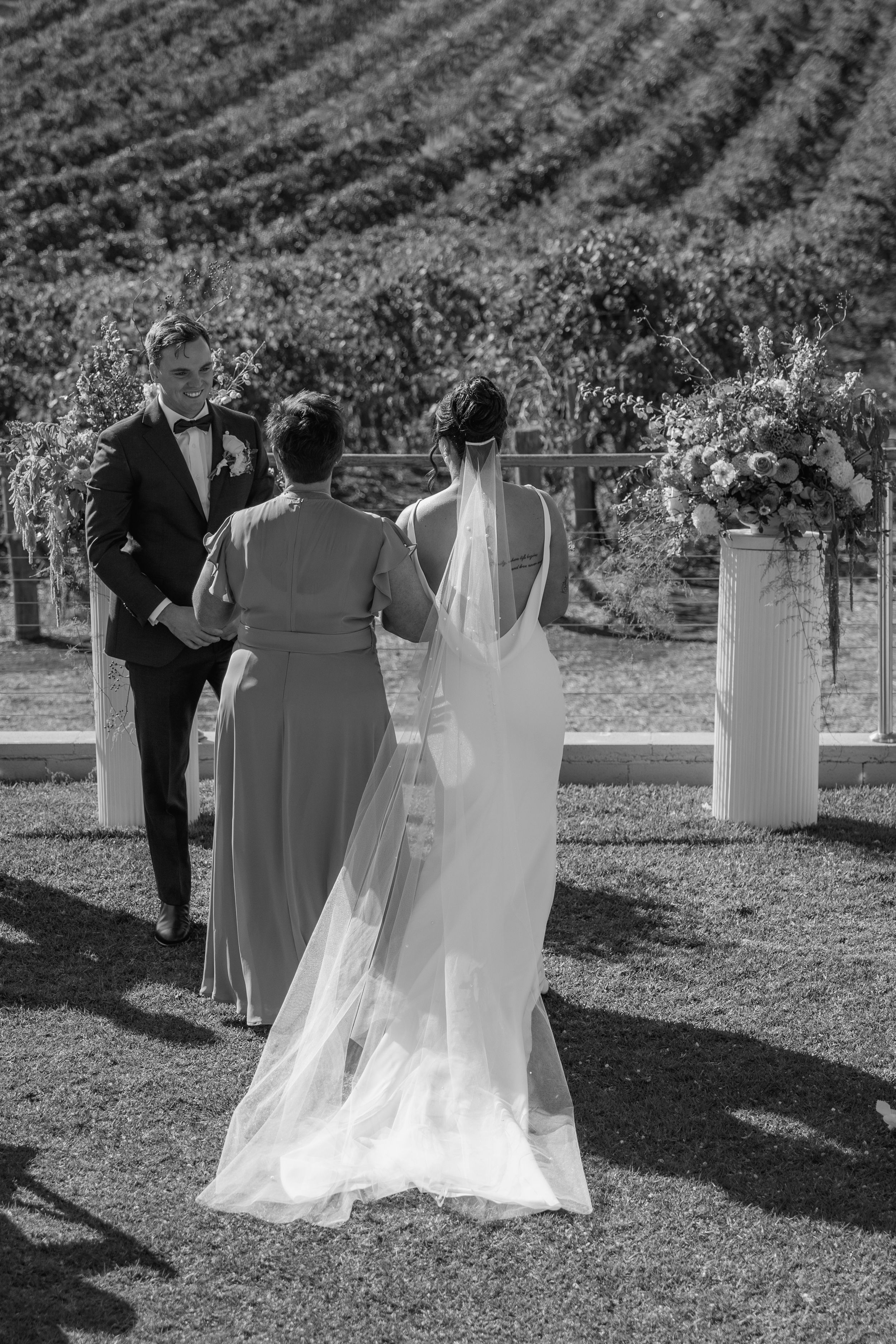 Wedding party members walk through vineyard rows during outdoor ceremony in black and white photograph.