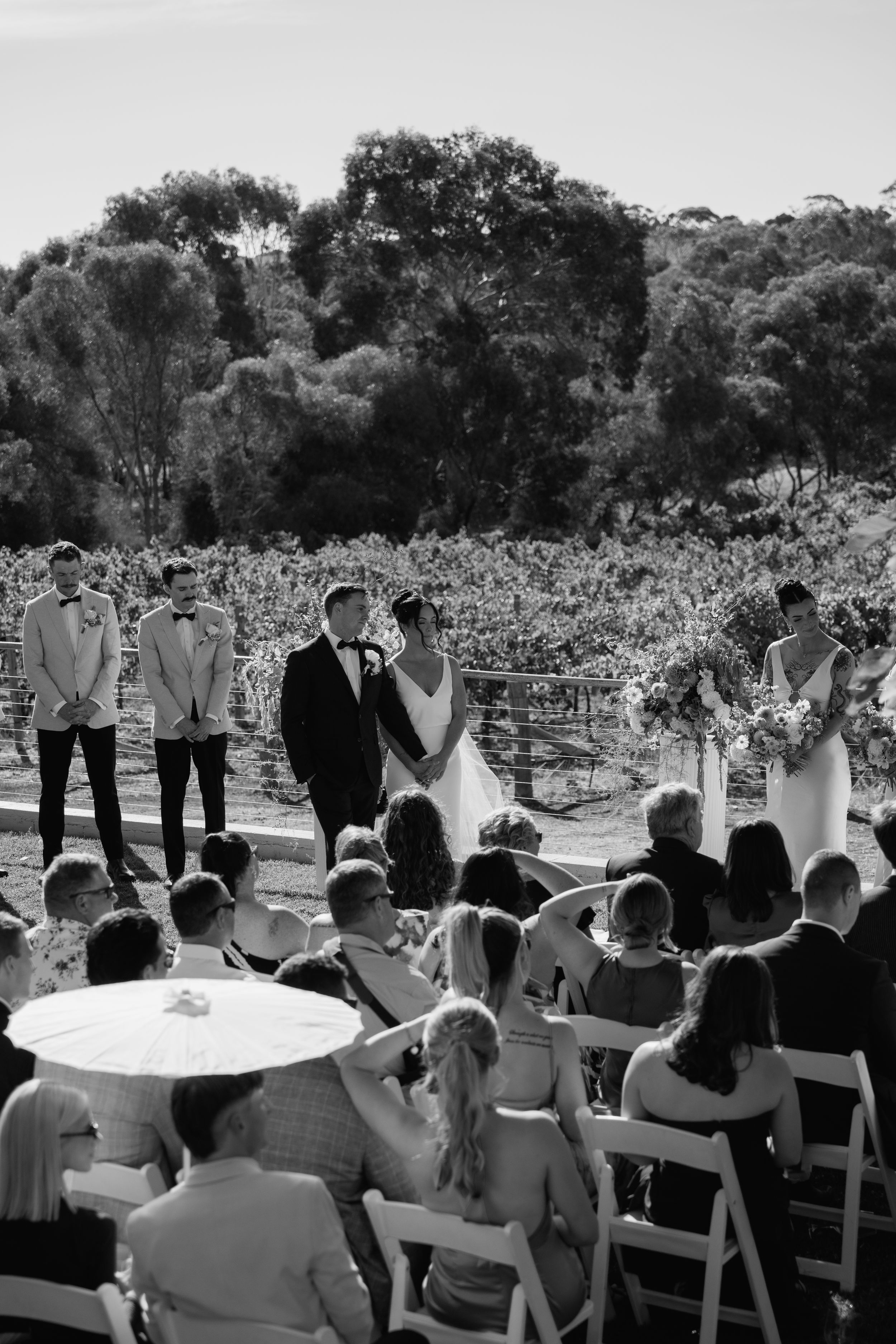 An outdoor wedding ceremony takes place in a vineyard with guests seated in white chairs watching the couple exchange vows.