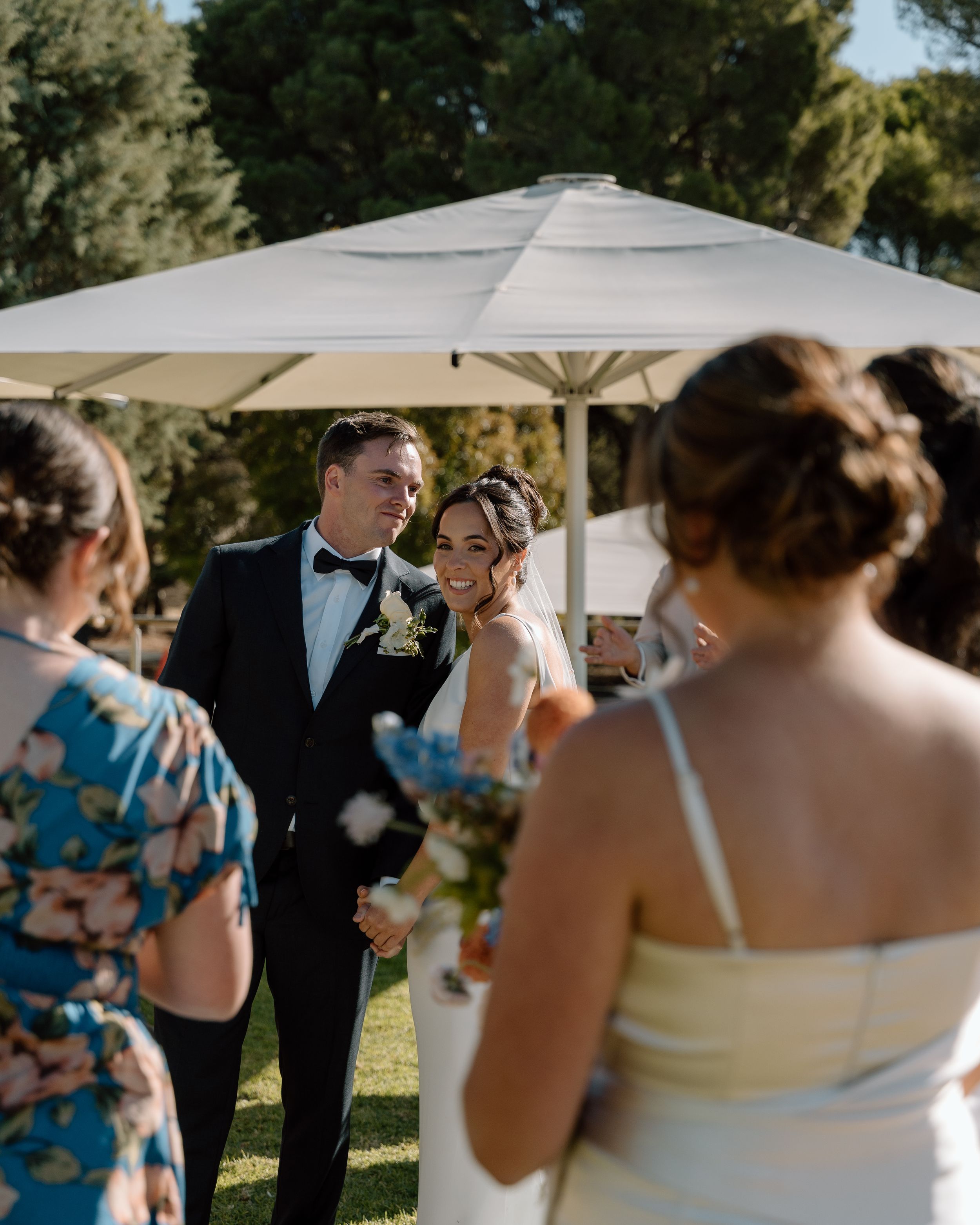 A wedding party celebrates outdoors under white umbrellas and trees on a sunny afternoon.