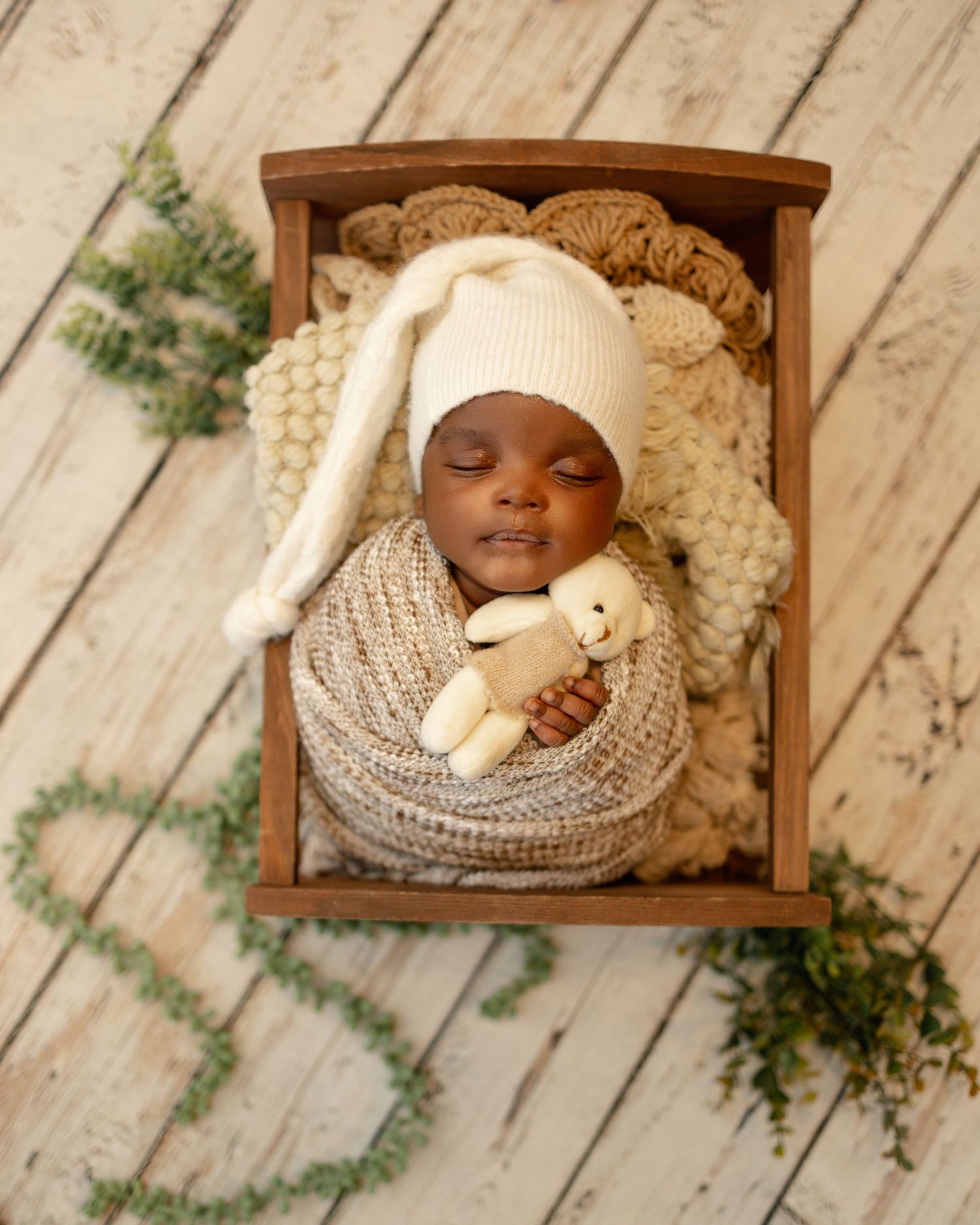 Sleeping baby swaddled in knit blanket wearing white bunny hat holds plush toy in wooden cradle with greenery decor.