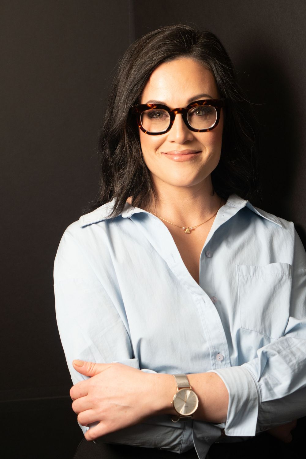 Professional headshot of woman in light blue button-down shirt and glasses against dark background.