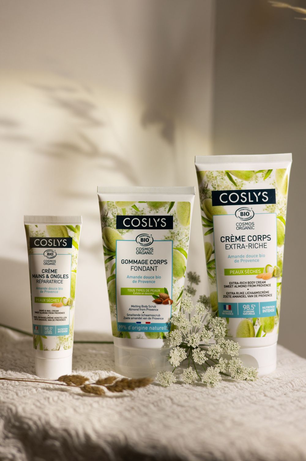 Trois produits de soins corporels biologiques Coslys disposés avec des fleurs séchées sur du tissu blanc.
