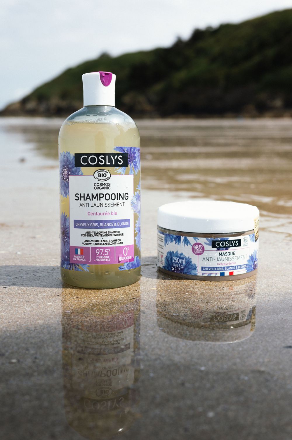 Shampooing biologique Coslys et produits de soins capillaires exposés sur le sable humide de la plage avec une toile de fond océanique.