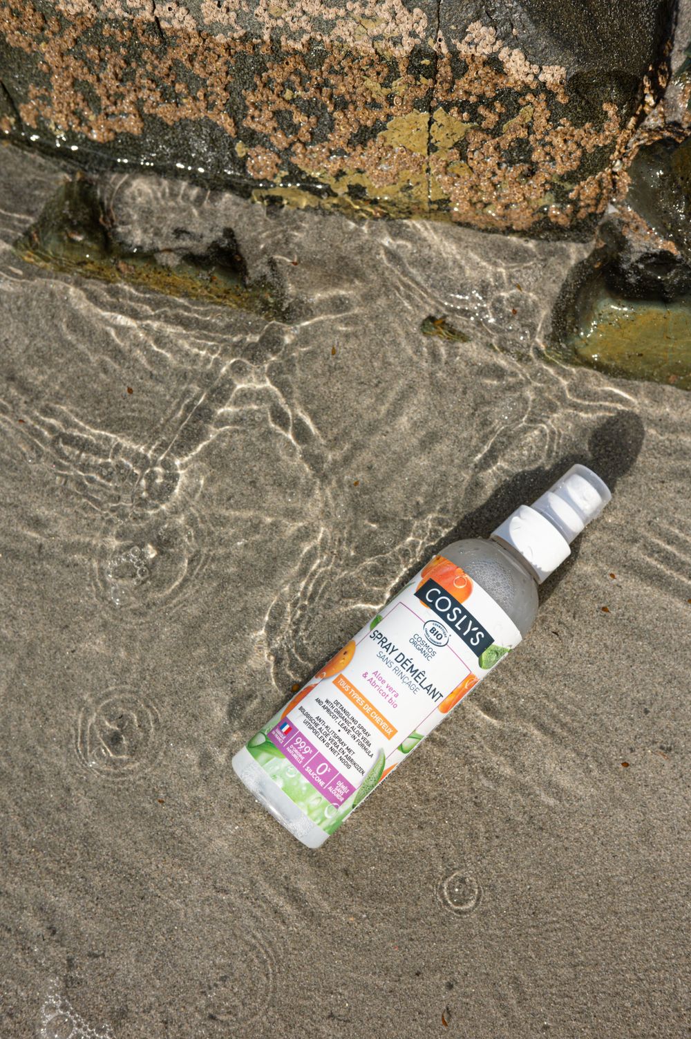 Bouteille de produit Coslys étiquetée orange posée sur une surface rocheuse texturée avec des motifs d'eau.