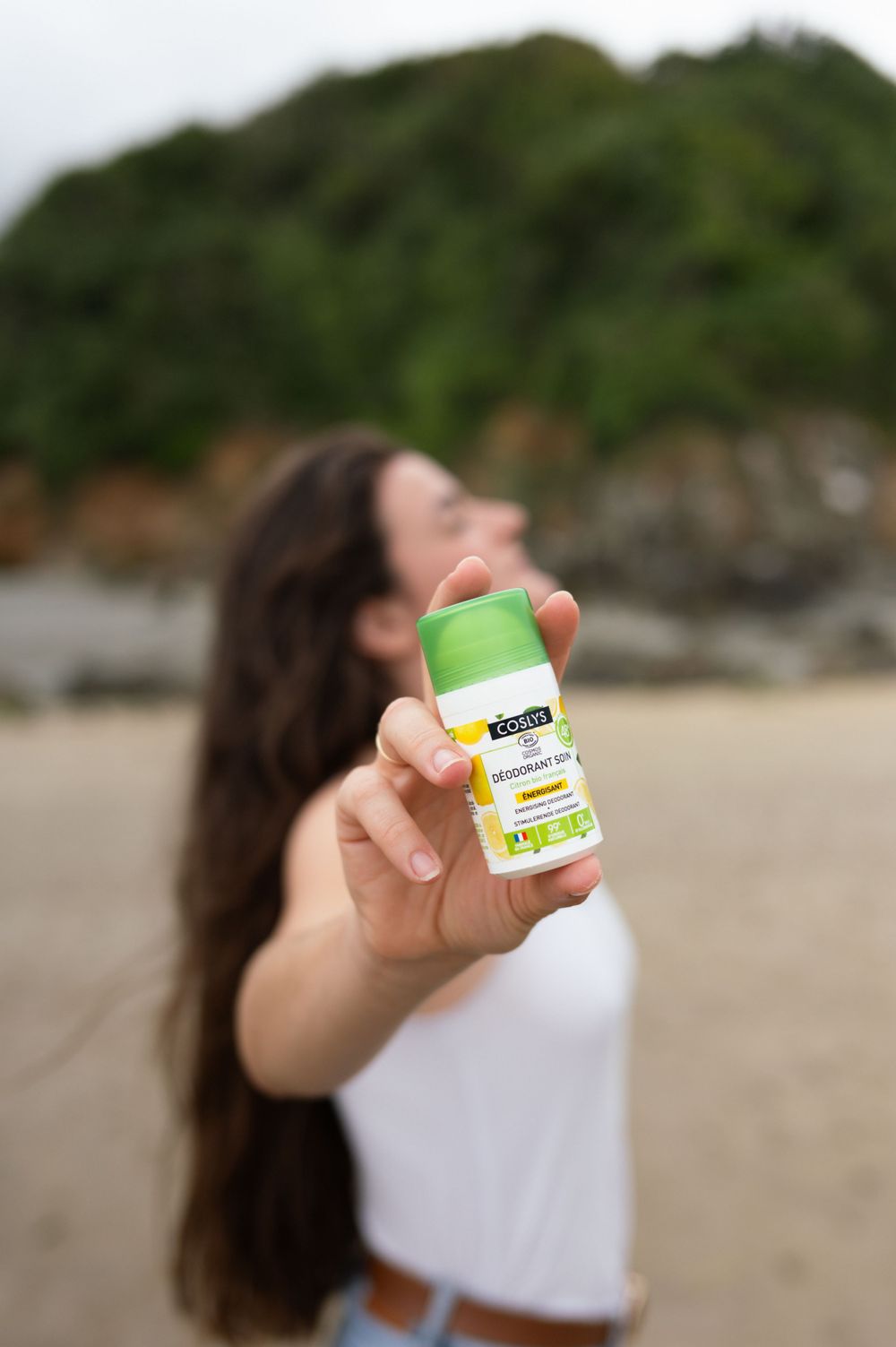 Main tenant une bouteille de produit naturel vert contre le paysage côtier flou.