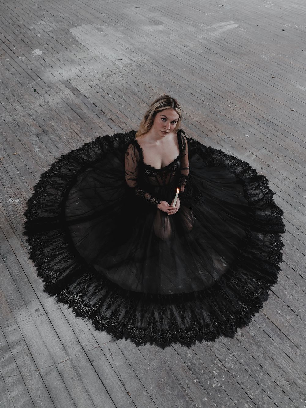 A woman in a dramatic black tulle gown with long sleeves spreads in a perfect circle on a wooden floor in moody lighting.