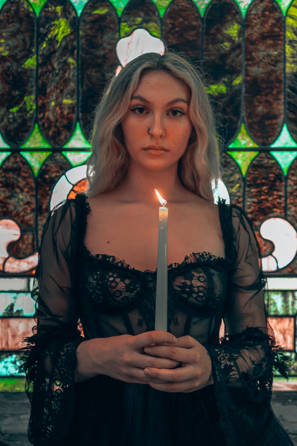 Woman in black lace dress holds lit white candle against stained glass window background in moody lighting.