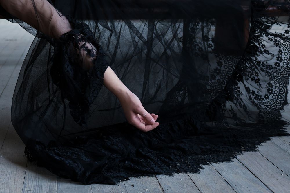 A woman's hand hanging down in black flowing dress lying on wooden bench in moody dark setting with dramatic lighting and shadows.