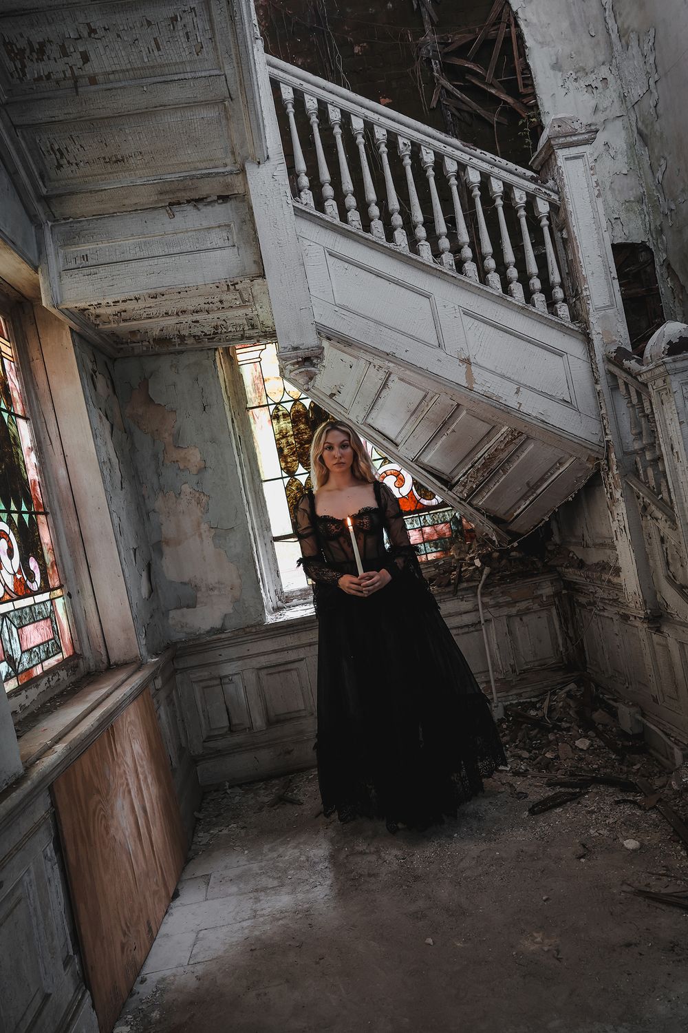 A woman holding a candle and standing in a deteriorating stairwell with peeling walls and debris on the floor.