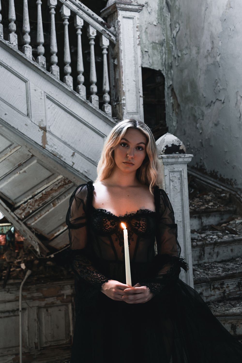 Atmospheric gothic scene of woman in black lace dress holding glowing candle on vintage staircase in abandoned building.