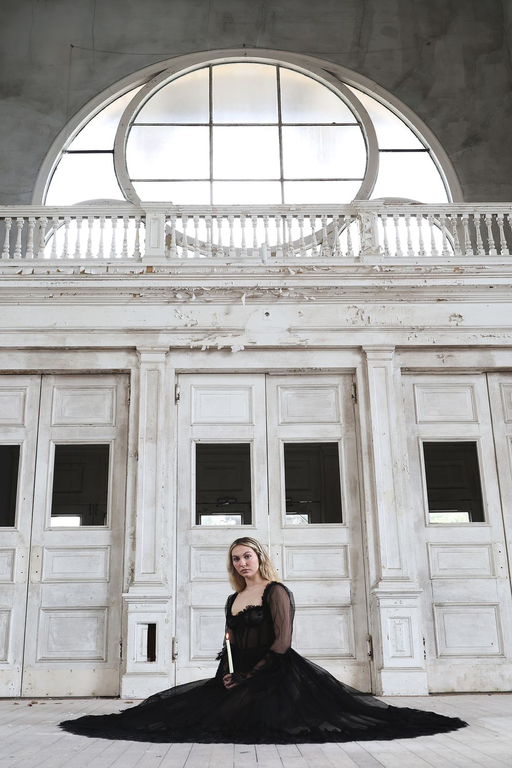 A woman in a black evening gown sits on the floor against ornate white doors beneath an arched window.