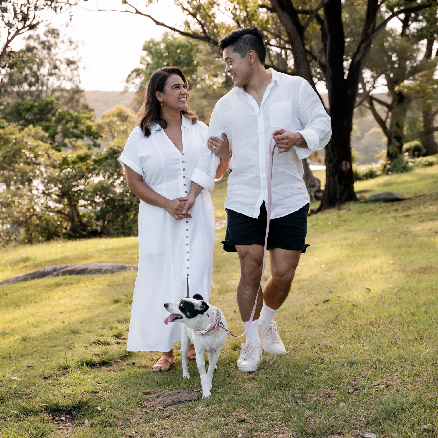 couple is laughing, looking at each other and walking towards the camera in the park while their dog is looking back at them on a photoshoot
