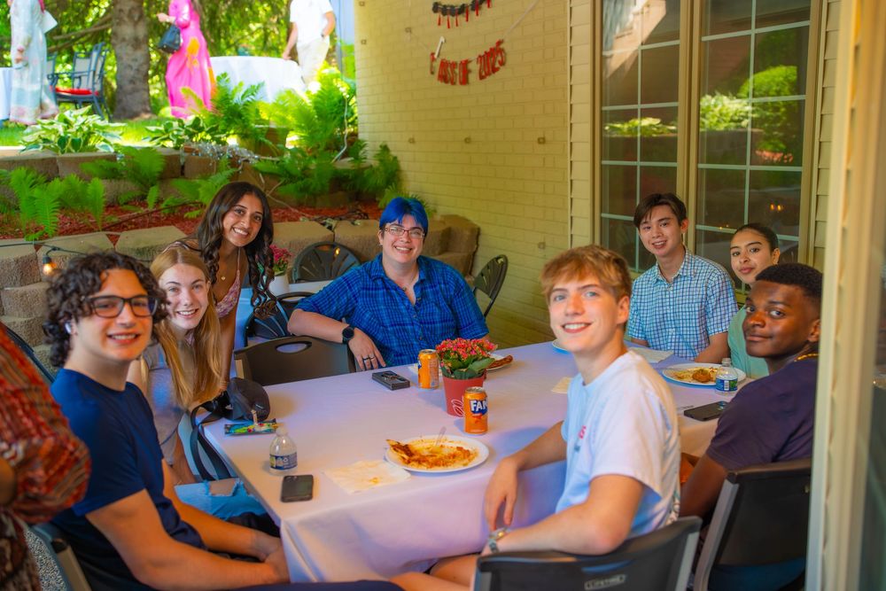 A group gathers around tables at an outdoor patio dining area surrounded by lush greenery and festive pink decorations. Quad Cities Grad