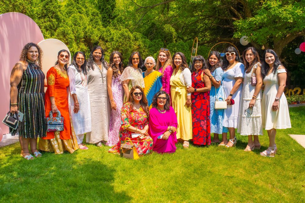 Large group photo at an outdoor garden party with pink wall backdrop and lush greenery.