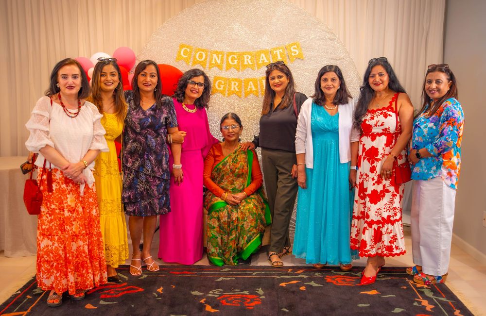 A group in brightly colored traditional dresses and formal wear stands in a line against a brown wall during an event.