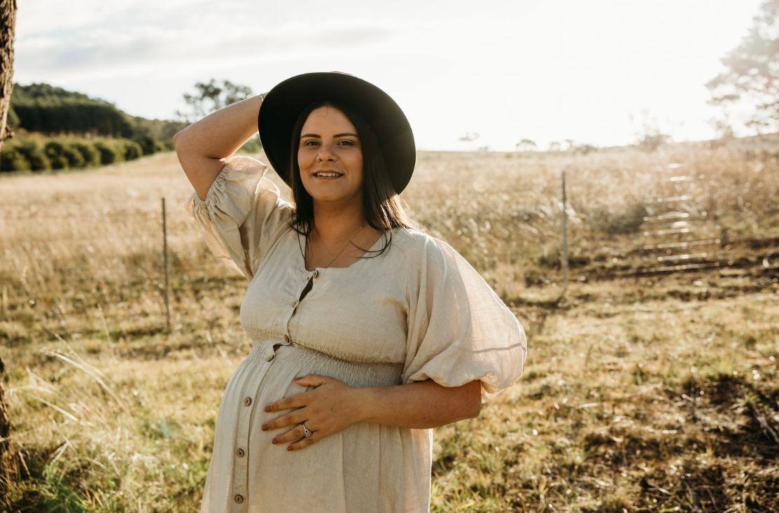 A person wearing a cream dress and black hat poses in a golden field during sunset, hand on belly.
