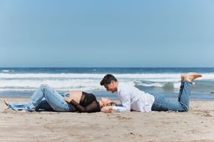 Couple lying on beach sand during maternity photo session with ocean waves in background.