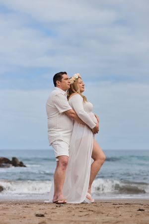 A couple in white attire sits together on a sandy beach with ocean waves and cloudy sky in the background.