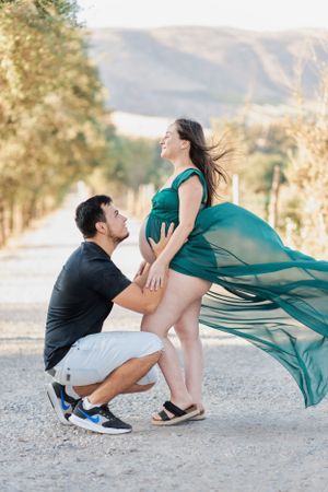 Expecting couple poses dramatically with flowing green dress in mountainous setting.