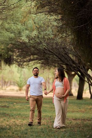 Expectant couple walks hand in hand through a park with overhanging trees during golden hour.