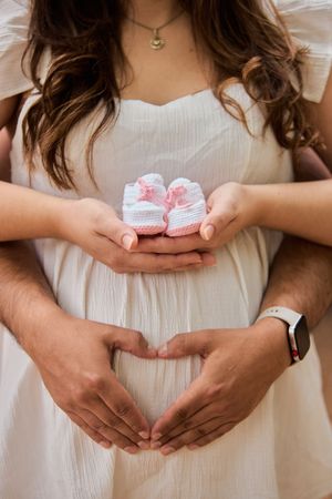 A series of photos showing hands holding tiny pink baby shoes against a white dress in a pregnancy announcement style.