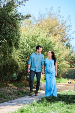A couple walks together on a garden path, with the person in a flowing blue dress showing signs of pregnancy.