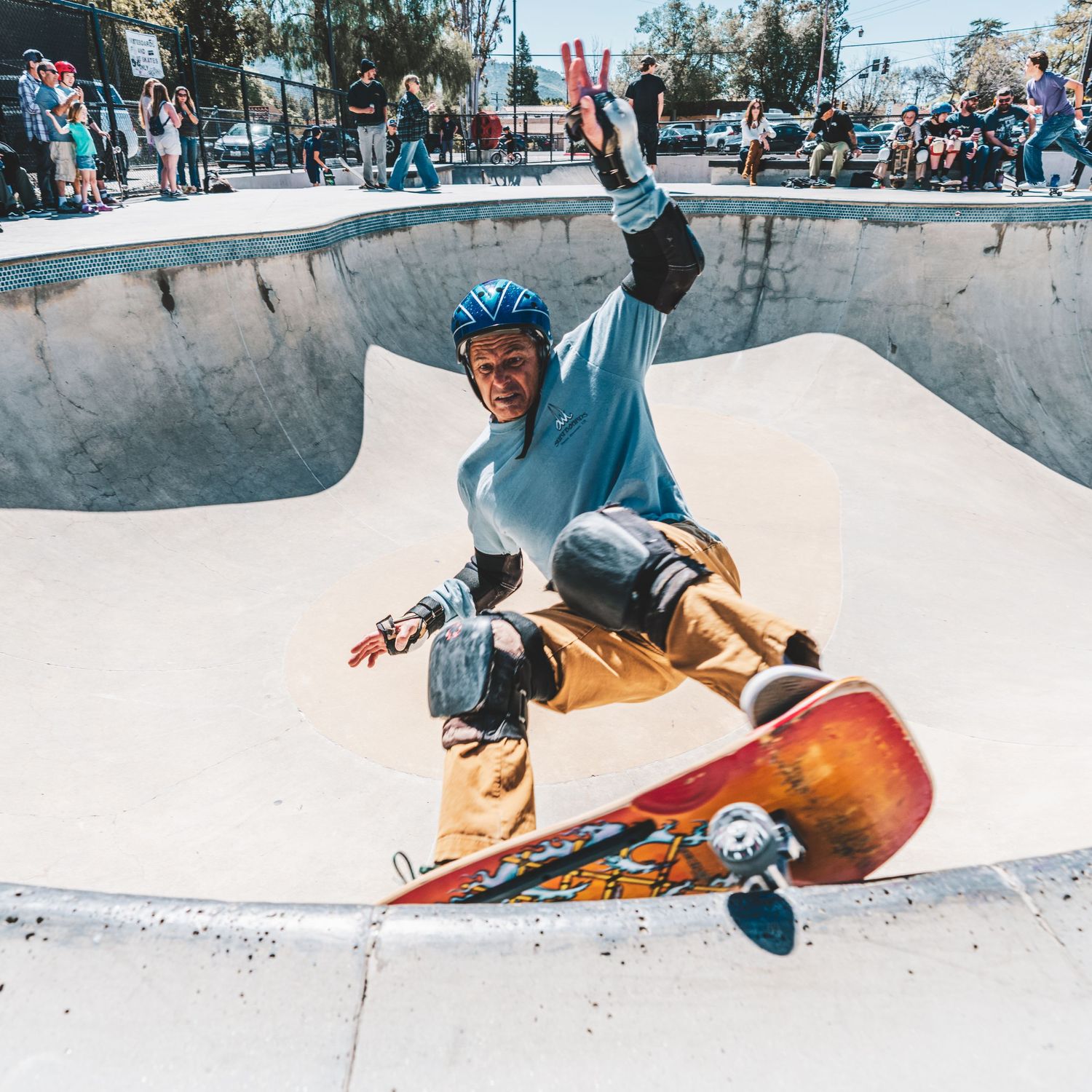 Skateboarder performs a trick in a concrete skate park bowl while wearing a light blue shirt and tan pants.