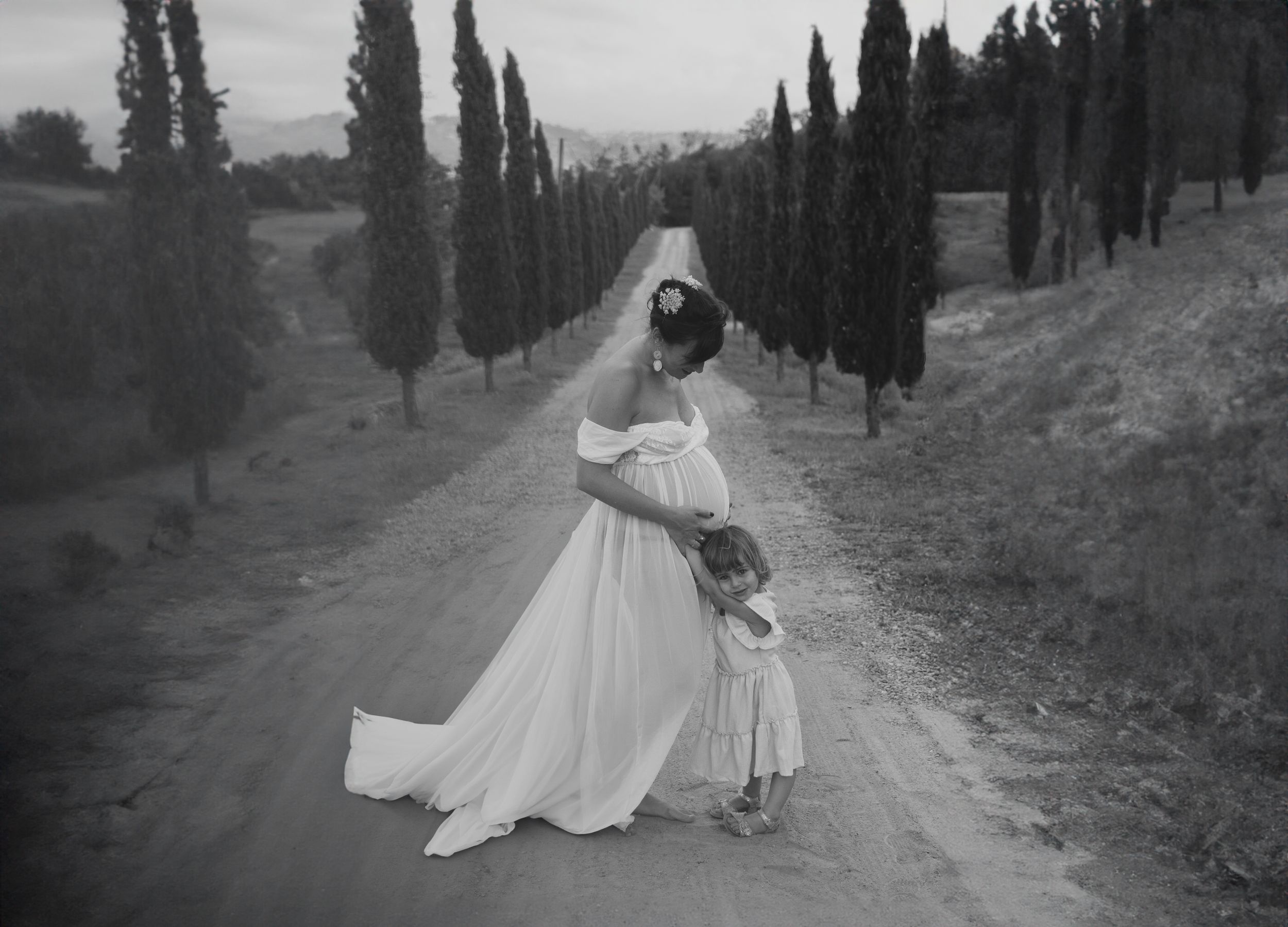 Foto in bianco e nero di madre in dolce attesa e figlia che abbraccia il pancione su una strada affiancata da cipressi. In Piemonte AL.