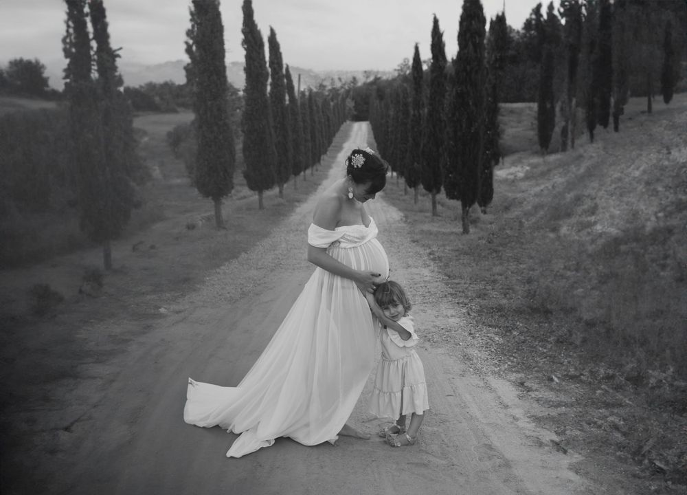 Foto in bianco e nero di madre in dolce attesa e figlia che abbraccia il pancione su una strada affiancata da cipressi. In Piemonte AL.