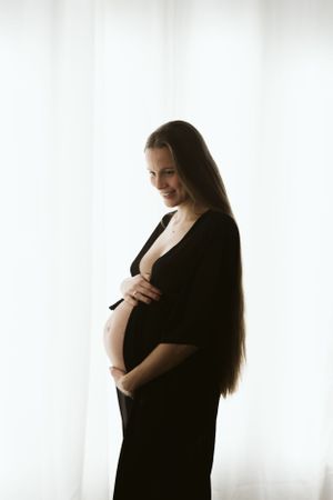 Profilo controluce una madre in dolce attesa in abito nero su finestra con tendaggio bianco. Servizio fotografico Maternità in provincia AL