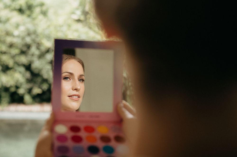 A reflection in a makeup mirror shows someone looking at themselves next to a colorful eyeshadow palette.