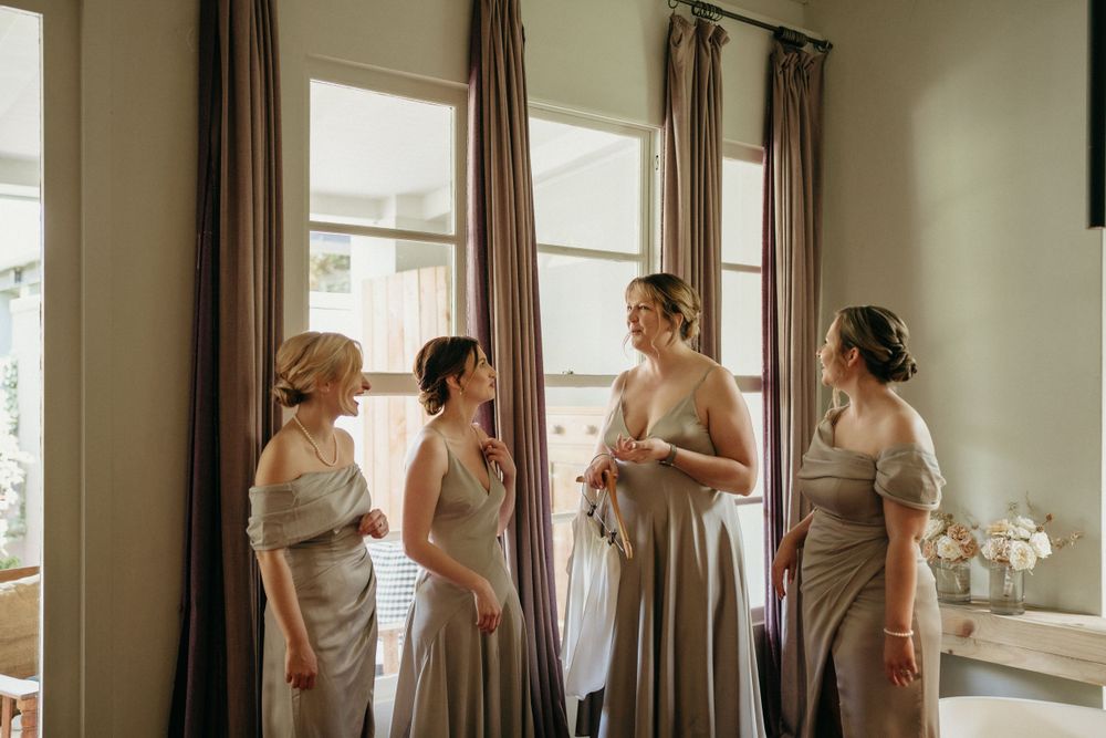 Bridesmaids in soft neutral-colored dresses gather by sunlit windows in an elegant getting ready room.