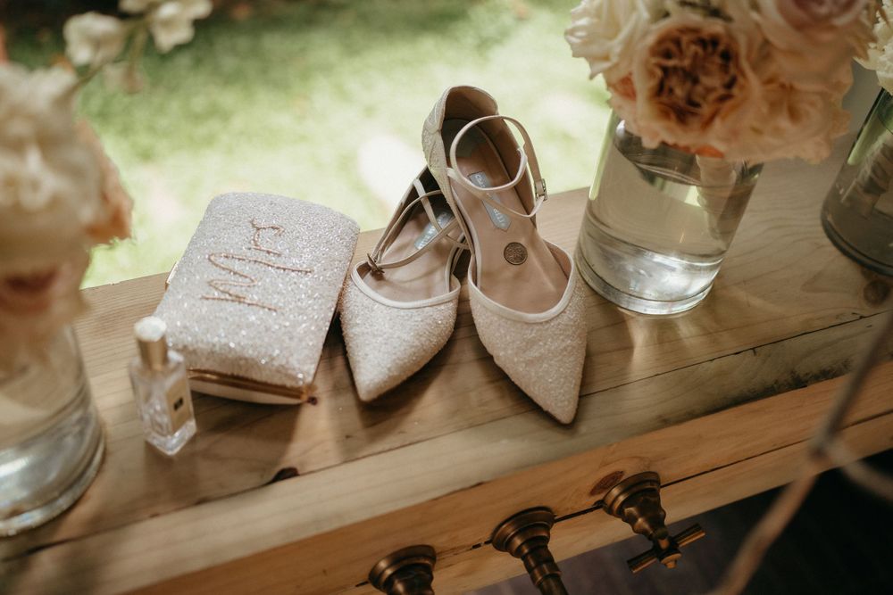 Delicate blush bridal shoes and wedding accessories arranged on wooden surface with floral decor.