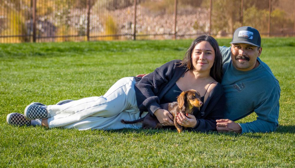 Smiling couple lying on green grass cuddling a dachshund puppy outdoors.