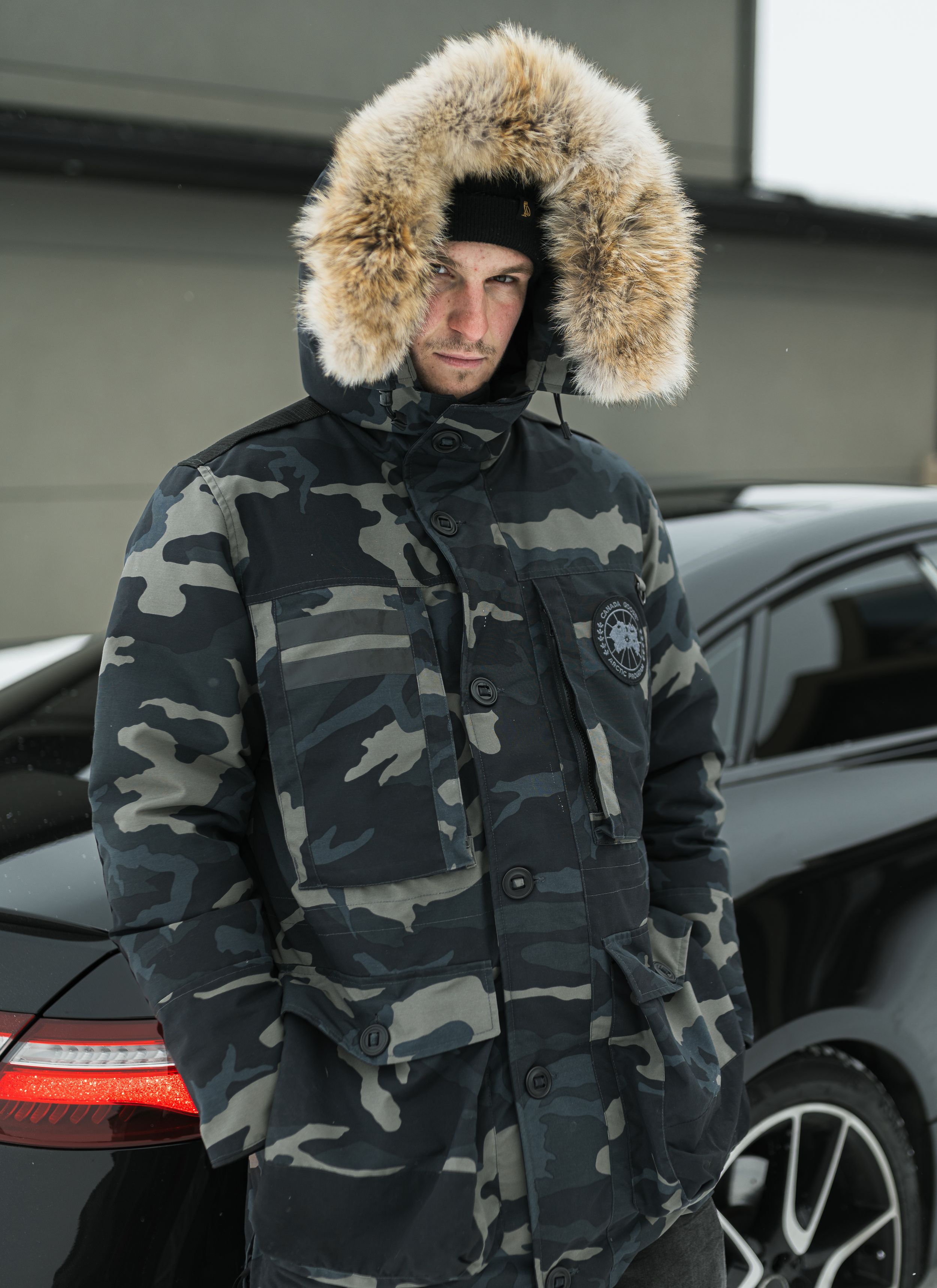 Person wearing a camouflage winter coat with fur-trimmed hood standing by a car.