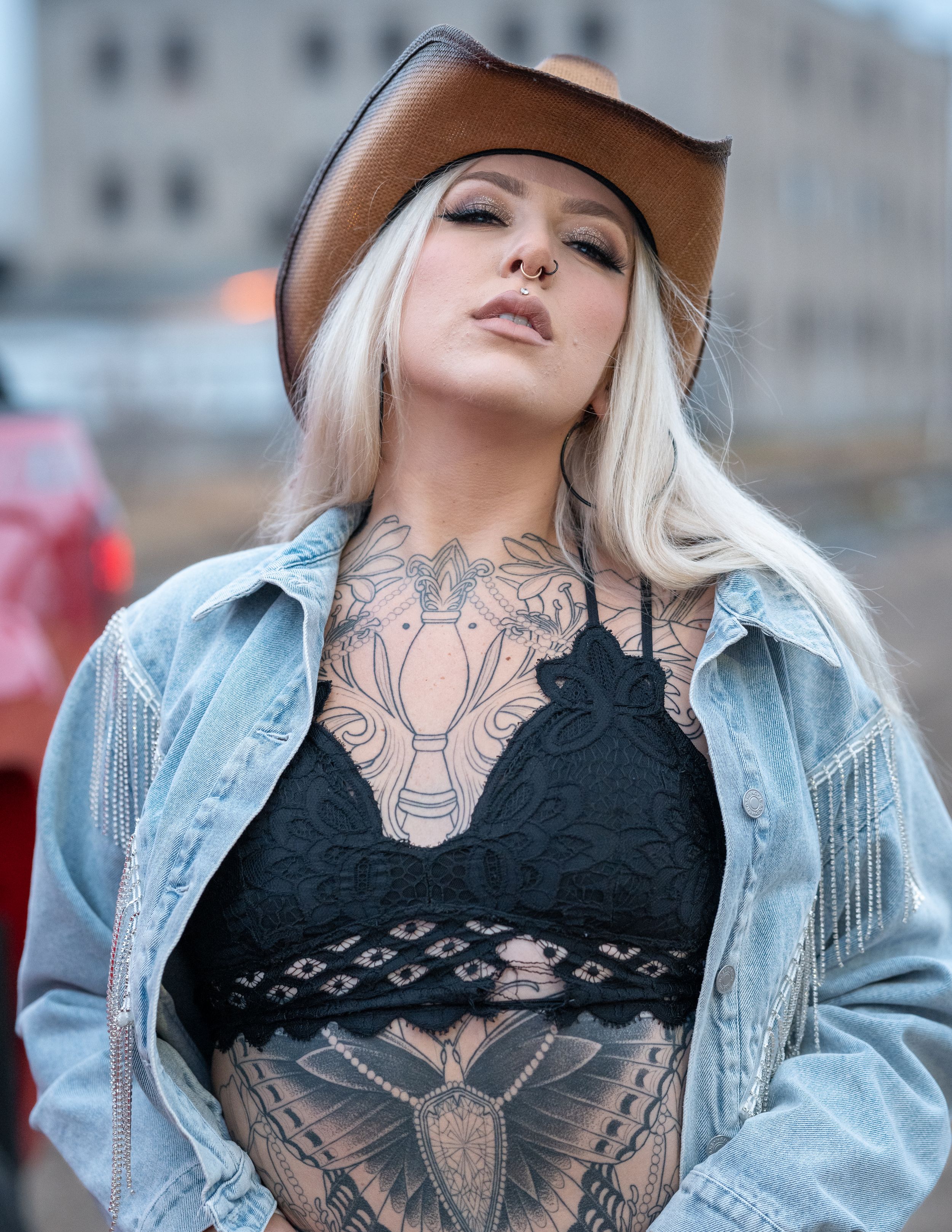 Stylish portrait showing platinum blonde hair, brown cowboy hat, denim jacket and visible tattoos in an urban setting.