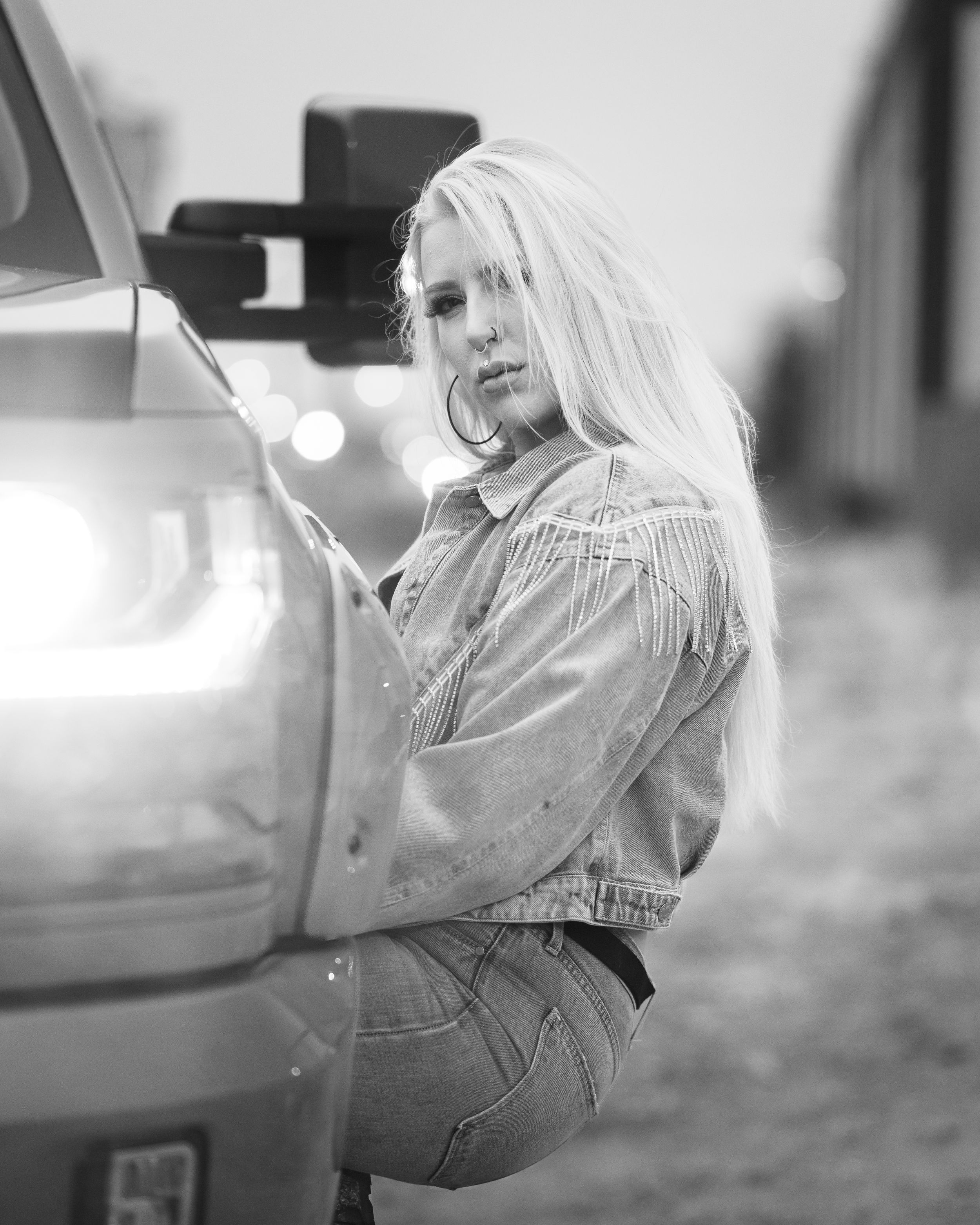 Black and white portrait of a person with long blonde hair leaning against a vehicle.