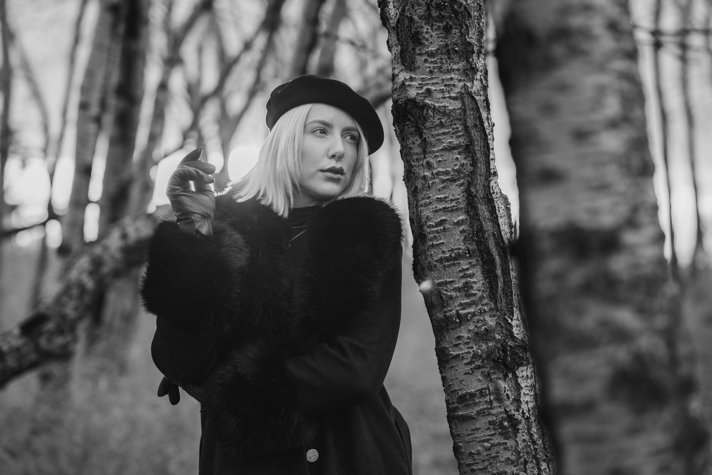 Black and white photo of someone in a dark sweater and beret leaning against a birch tree in a forest.