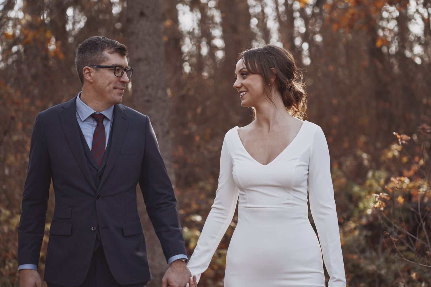 A newlywed couple walks hand in hand through an autumn forest, sharing a loving gaze as golden leaves frame their intimate moment.