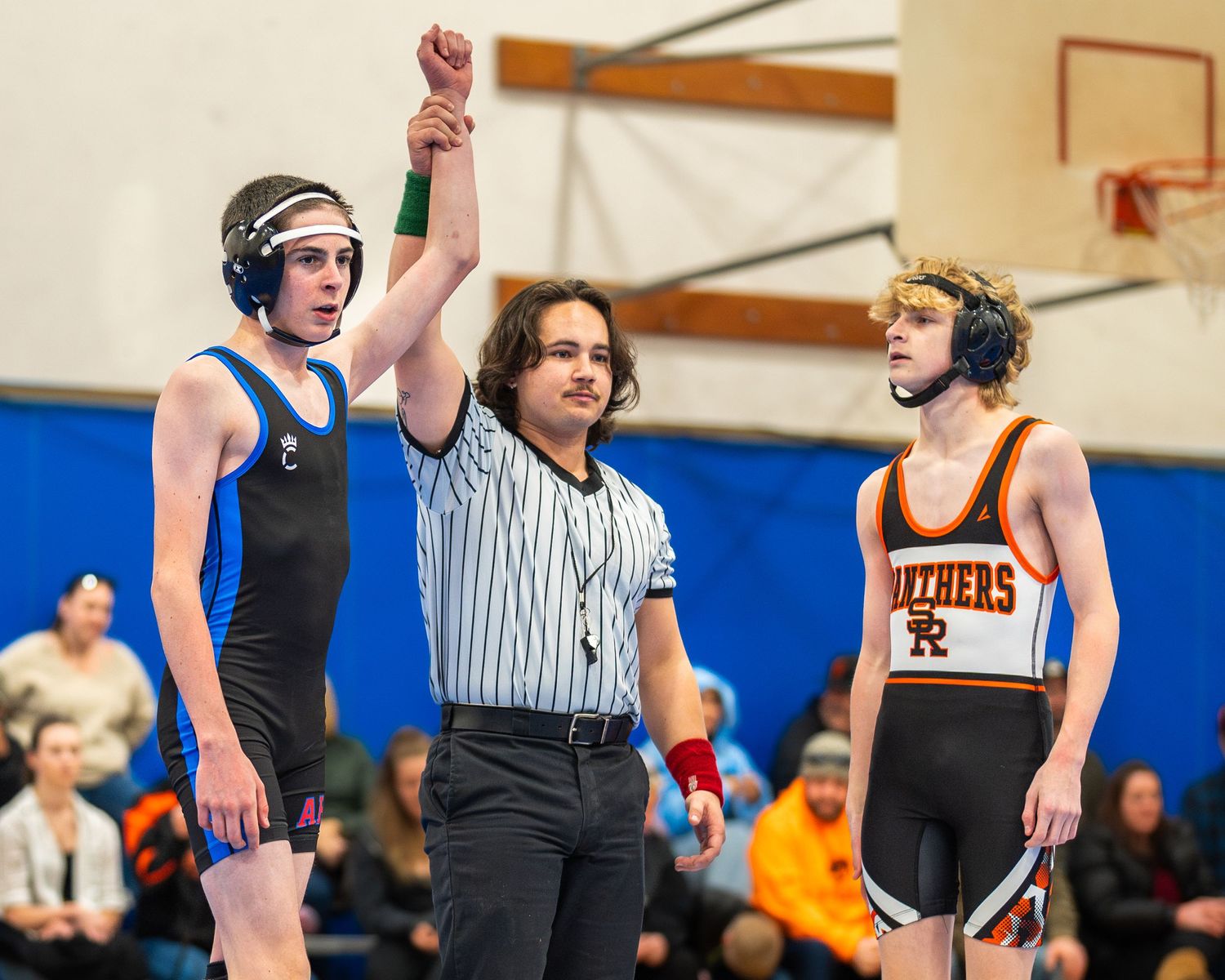 A wrestling referee raises a competitor's arm in victory during a high school match while another wrestler looks on.