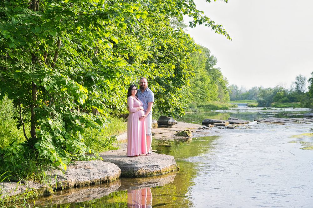 Two people in flowing gowns stand on rocks by a tranquil river surrounded by lush summer greenery.