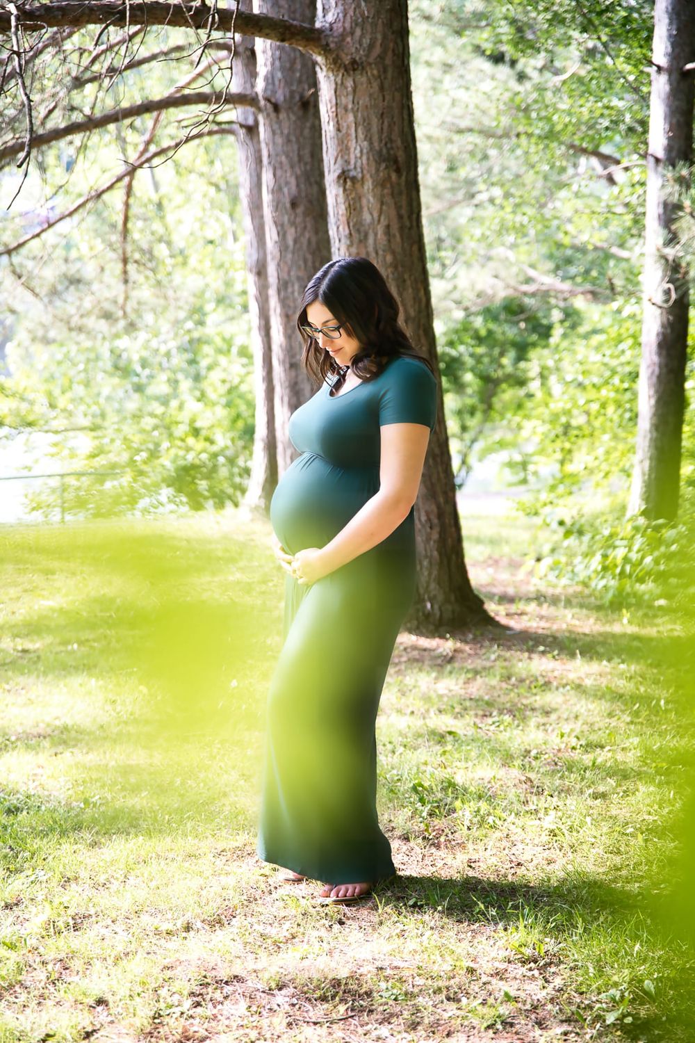 Expectant mother in a teal dress stands peacefully under tall pine trees in a sunlit forest setting.