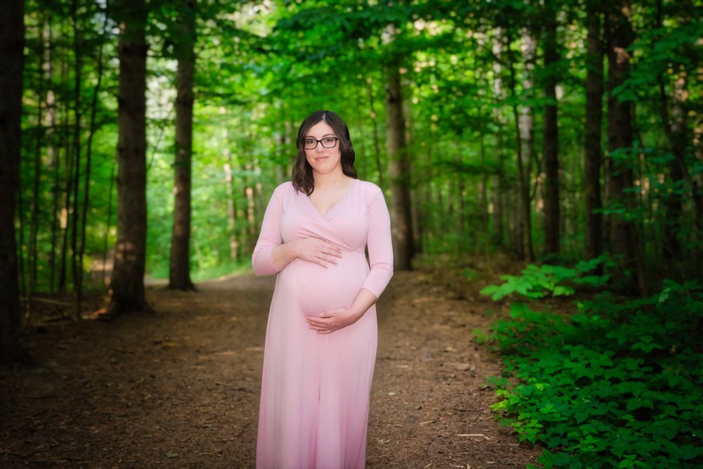 Elegant maternity portrait of woman in flowing pink dress standing on forest path surrounded by lush greenery.