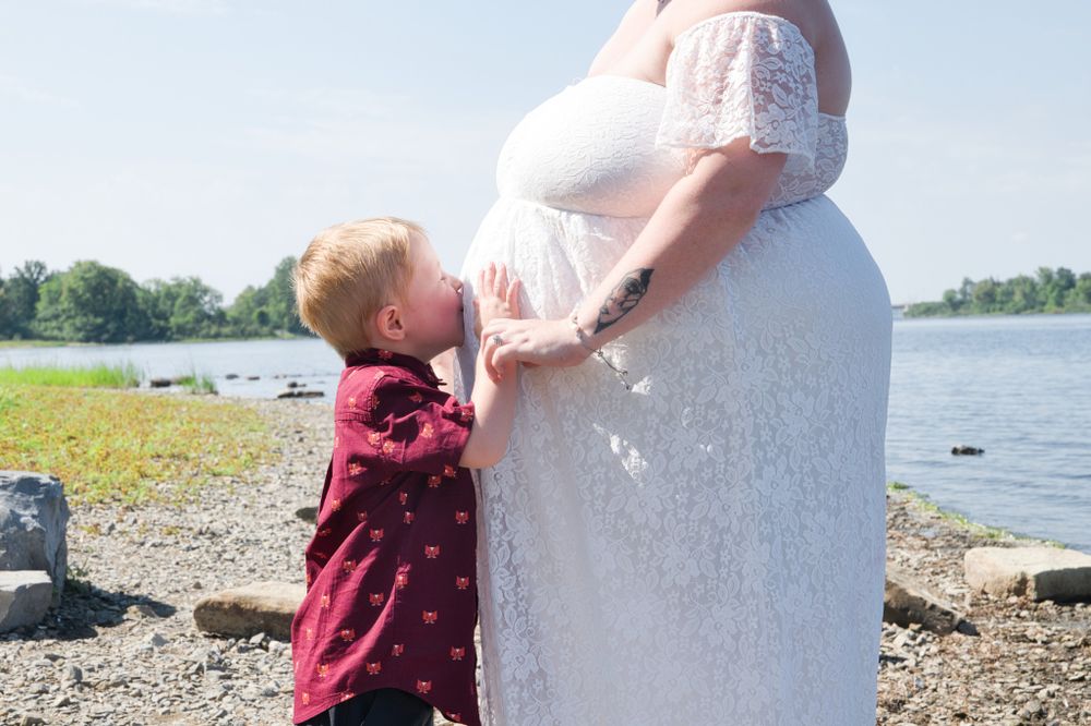 Small child touching a pregnant person's belly on a lakeside beach.