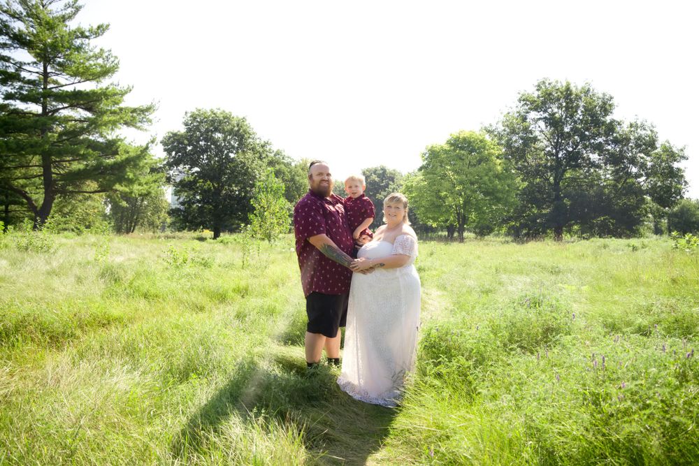 A couple in outdoor wedding attire pose together in a sunny summer meadow with trees in the background.