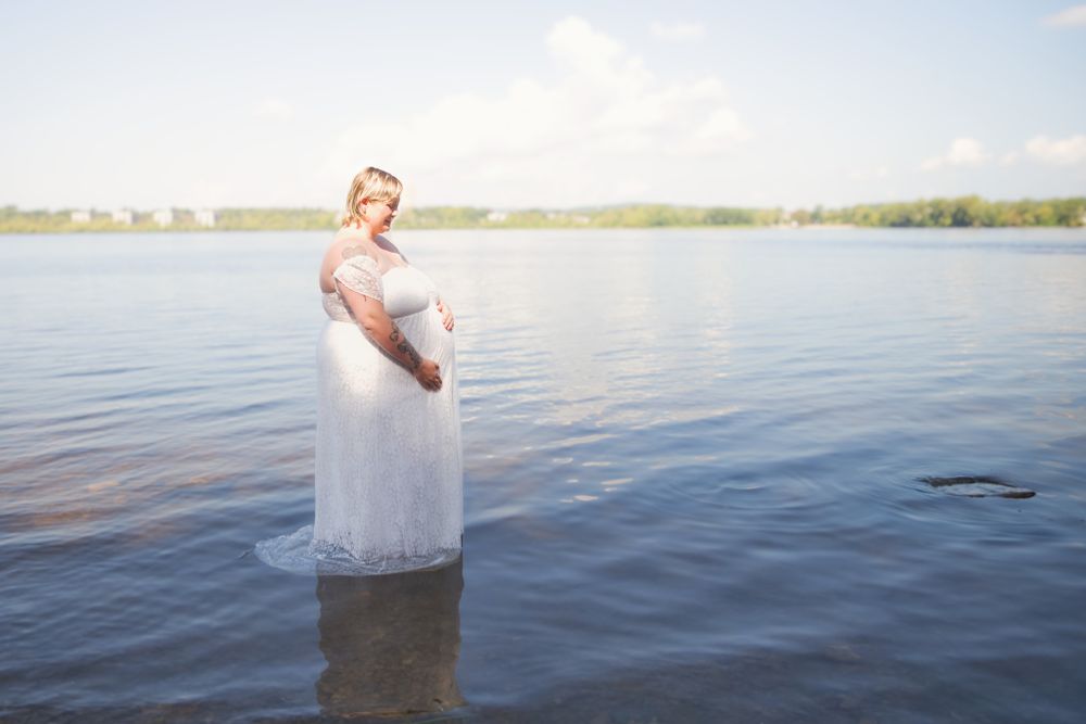 A person in a white dress stands in calm lake waters during a sunlit outdoor photo session.