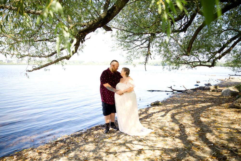 A romantic couple embraces on a scenic lakeshore under overhanging tree branches.