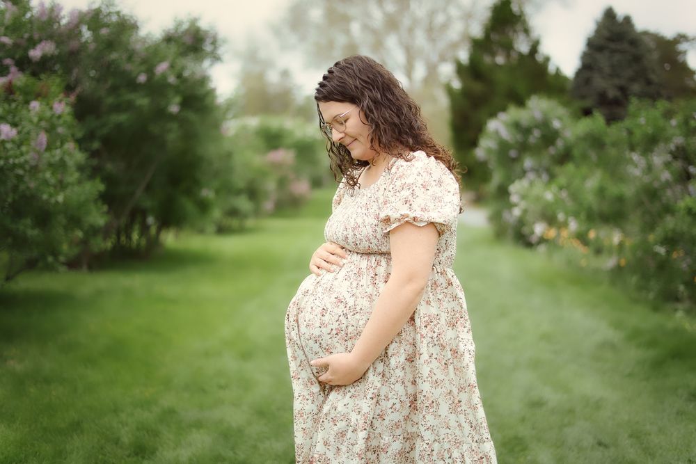 A pregnant woman in a floral dress stands in a lush garden setting during a maternity photoshoot.