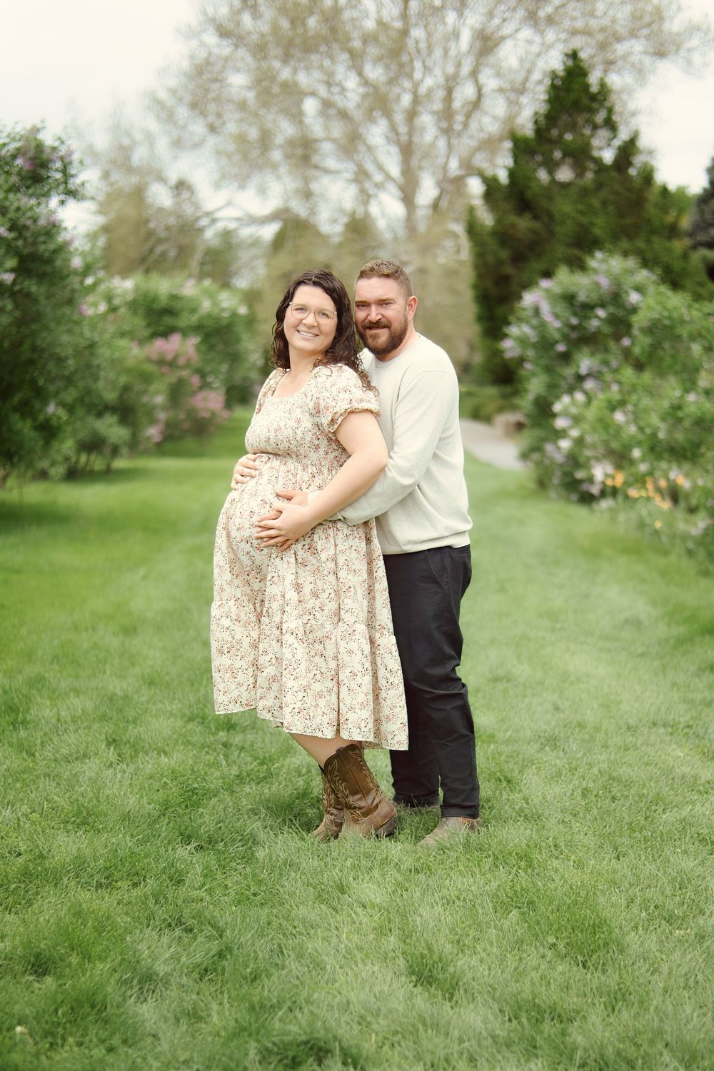 A couple embraces on lush green grass in a garden setting wearing floral dress and white shirt.