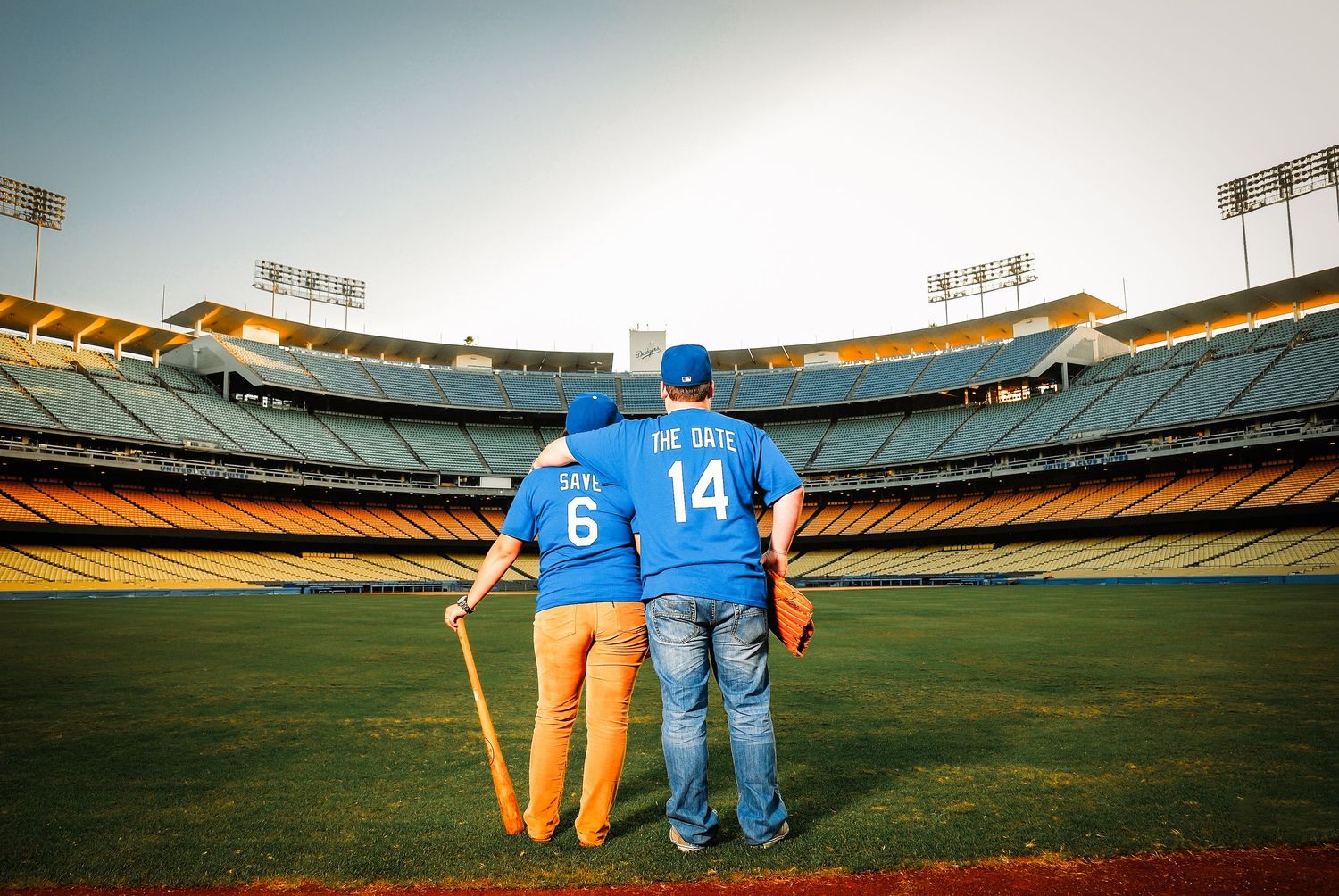 Two baseball players in numbered jerseys stand together on an empty field inside a large stadium during daytime.